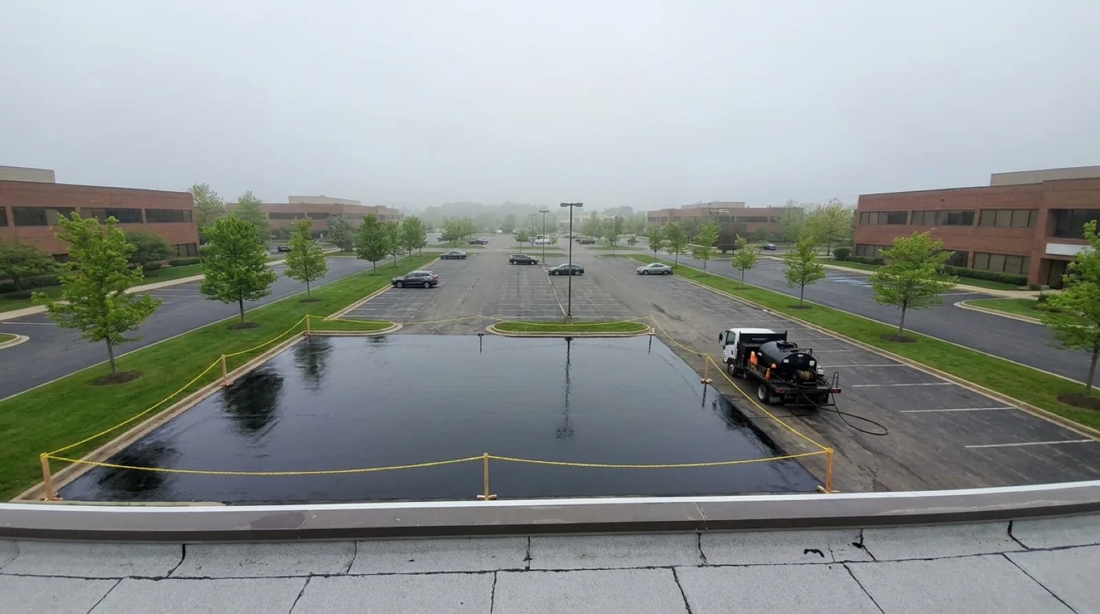 An elevated view of a business-park lot where one roped-off section is freshly sealcoated beside a small sealer truck while cars park farther away.