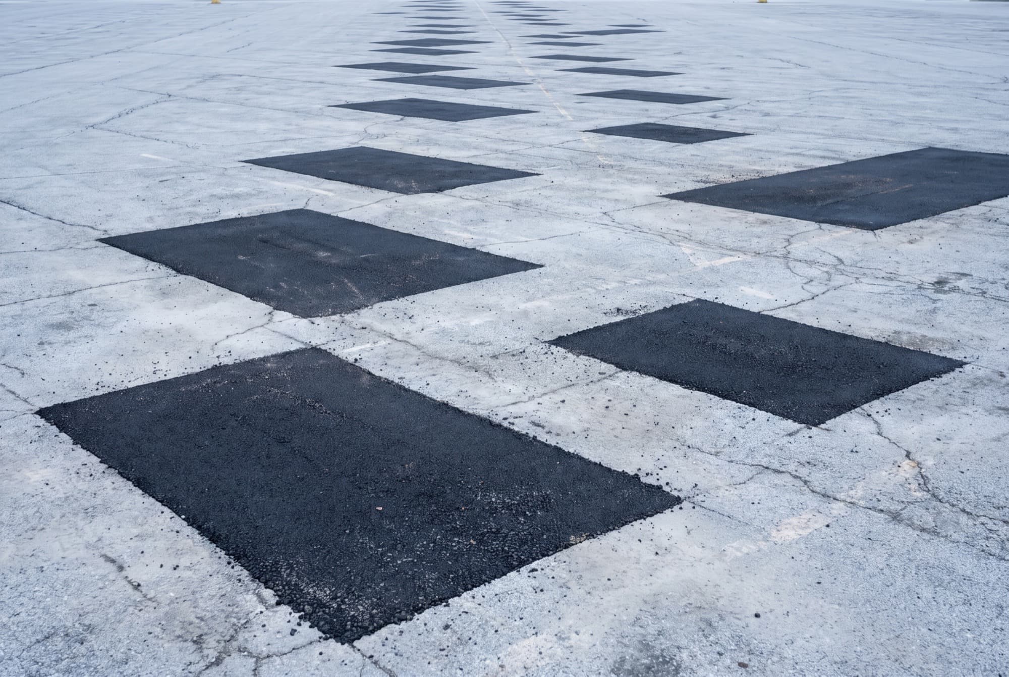 The surface of a commercial parking lot in weathered pale grey asphalt with a scattering of rectangular saw-cut repair patches set into it, each patch a clean square of new black asphalt with hard straight edges against the cracked older surface.