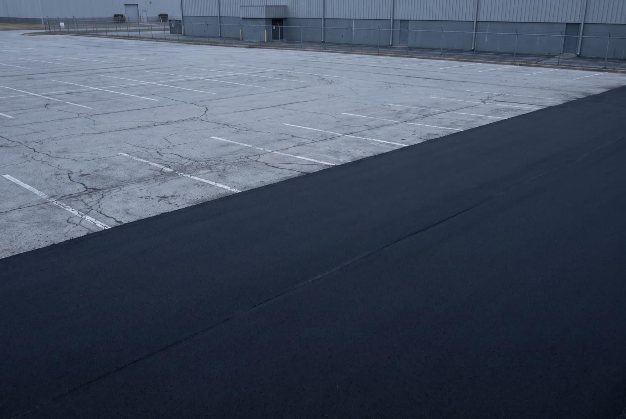 A commercial parking lot part way through resurfacing. The near half is uniformly new black asphalt with no lines on it yet, the far half is the old cracked pale grey surface it replaces, and a straight sharp joint runs between the two.
