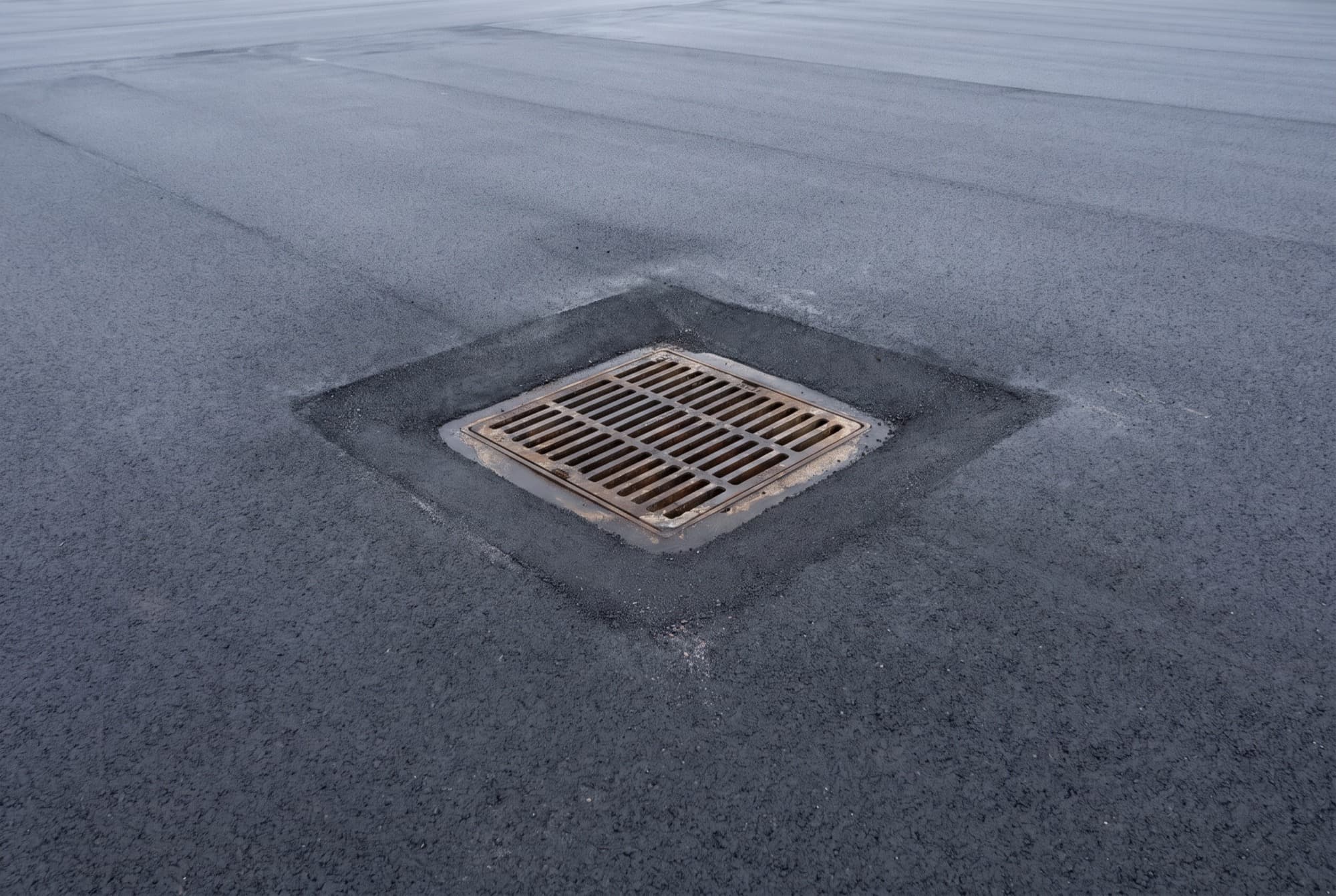 A low point of an empty commercial asphalt parking lot seen from above, where a square cast iron catch basin grate sits flush in its frame in new black asphalt, with the surface sloping gently in toward it from every side.