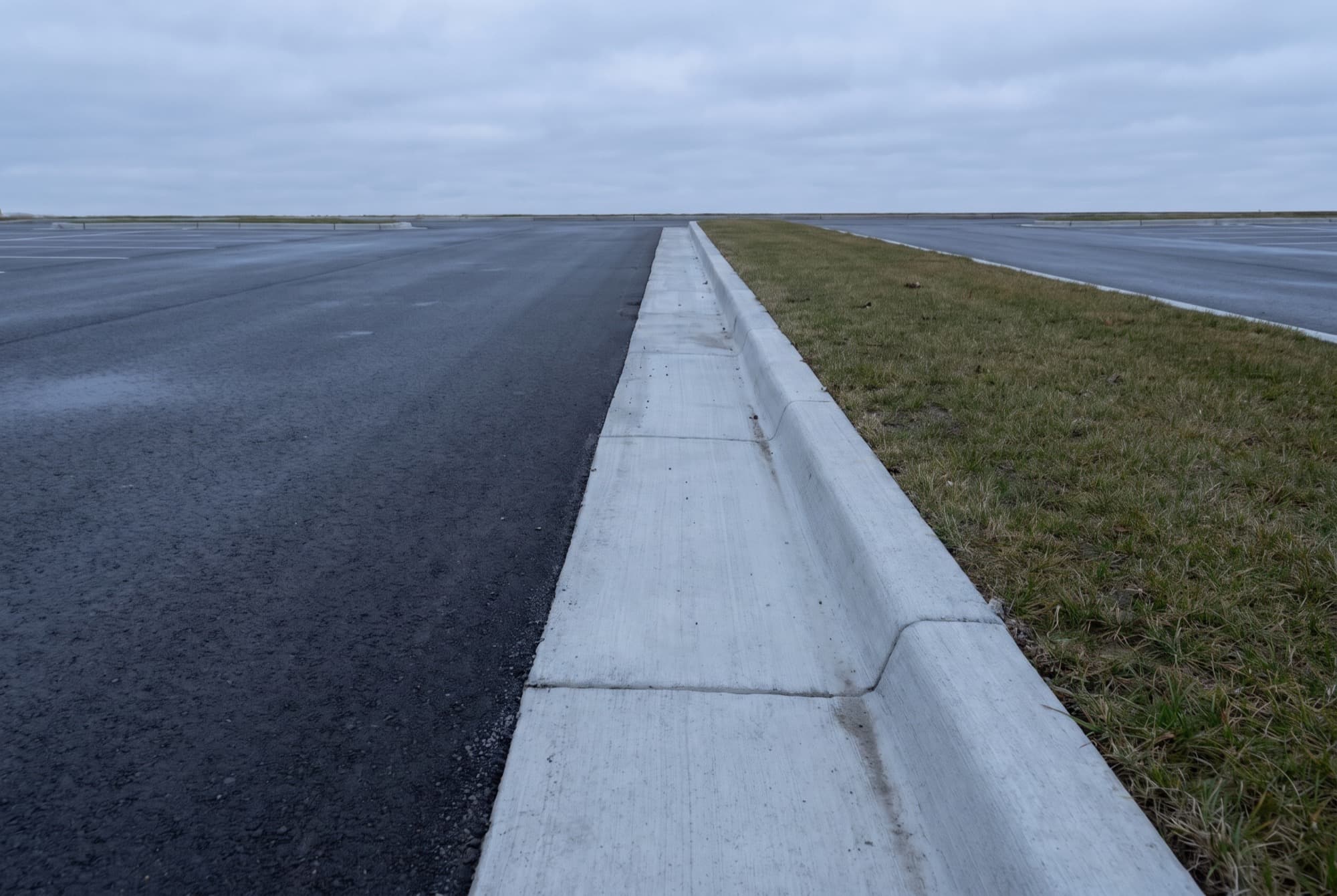 The edge of an empty commercial asphalt parking lot where a new pale grey concrete curb and gutter runs away into the distance as one continuous formed unit, black asphalt on one side of it and a plain grass verge on the other.