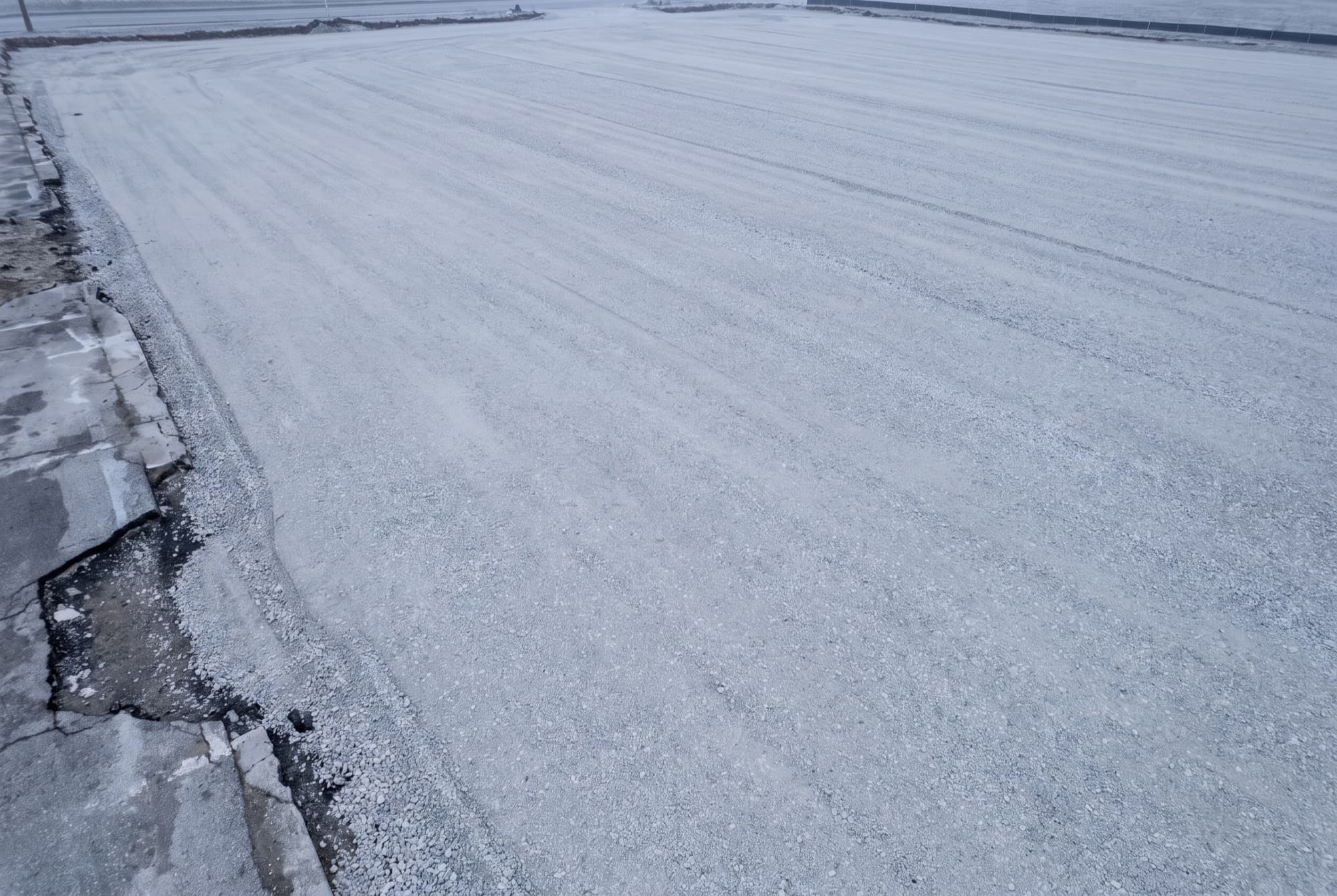 A commercial lot stripped back to bare compacted granular base, a flat expanse of pale grey crushed stone graded to a shallow even fall, with no asphalt anywhere and the cut edge of the removed pavement visible along one side.