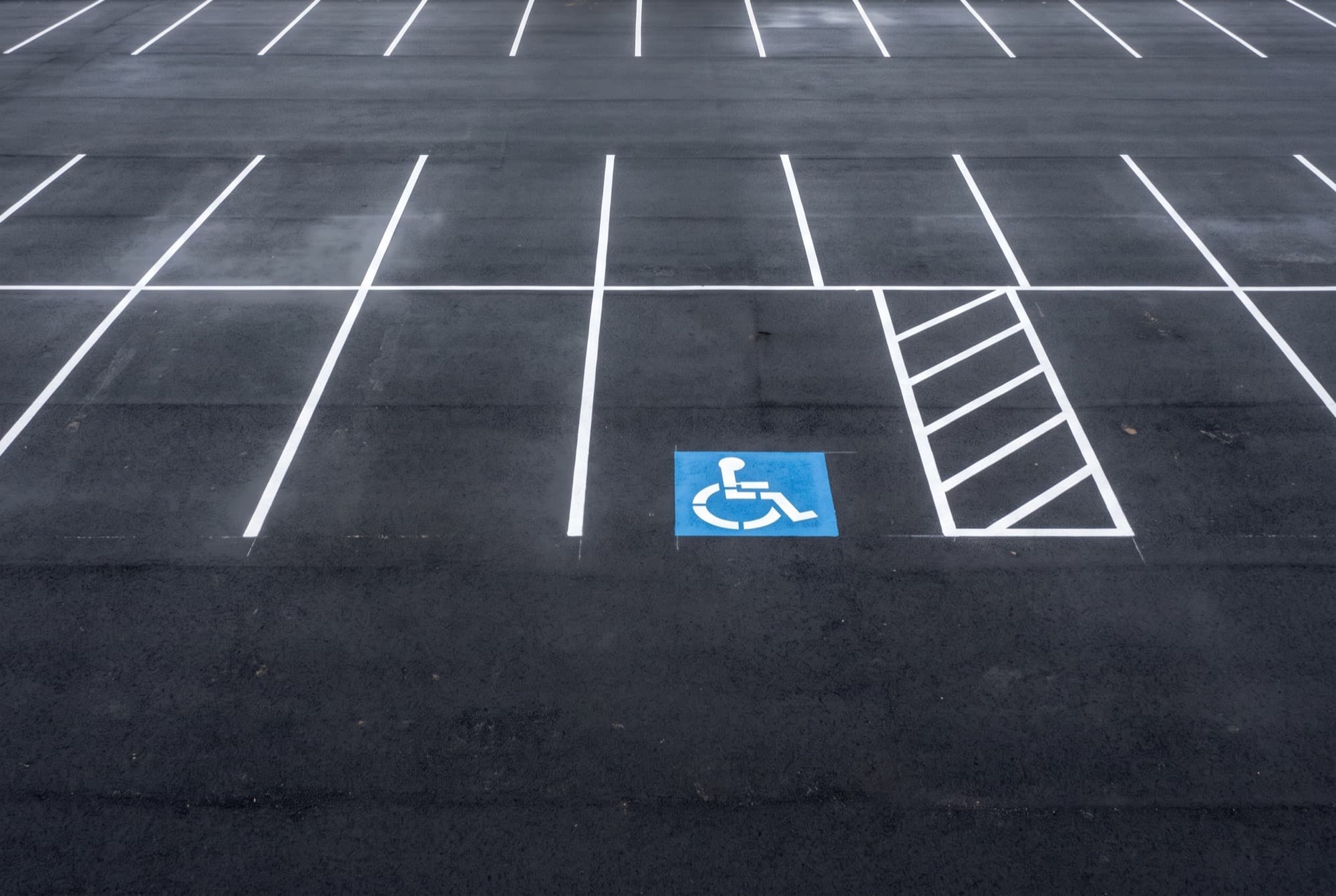 A section of an empty asphalt parking lot seen almost from above, filled with fresh crisp white stall lines on black asphalt, one blue and white painted accessible parking symbol, and a hatched access aisle of white diagonal stripes beside it.