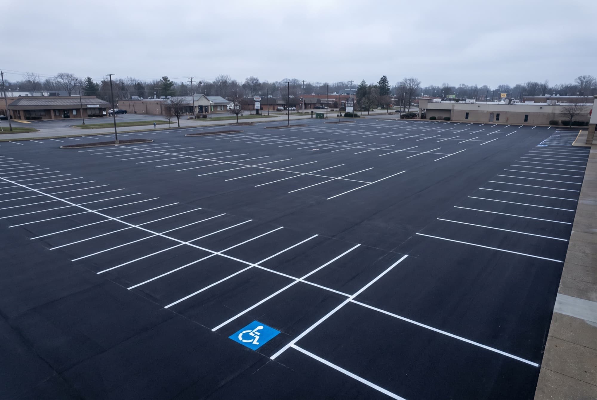 An empty commercial plaza parking lot the morning after it was paved and lined. The asphalt is uniformly new and black, the stall lines are freshly painted white, and one blue and white accessible parking symbol is the only colour in the frame.