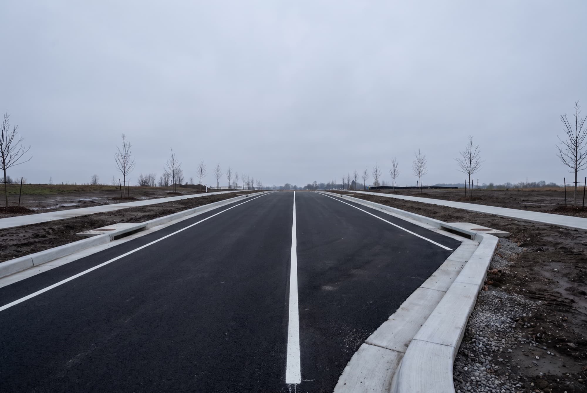 A newly built empty two lane municipal road, the asphalt uniformly new and black, a crisp white centre line and white edge lines running away to the horizon, with new pale grey concrete curb and gutter along both sides.
