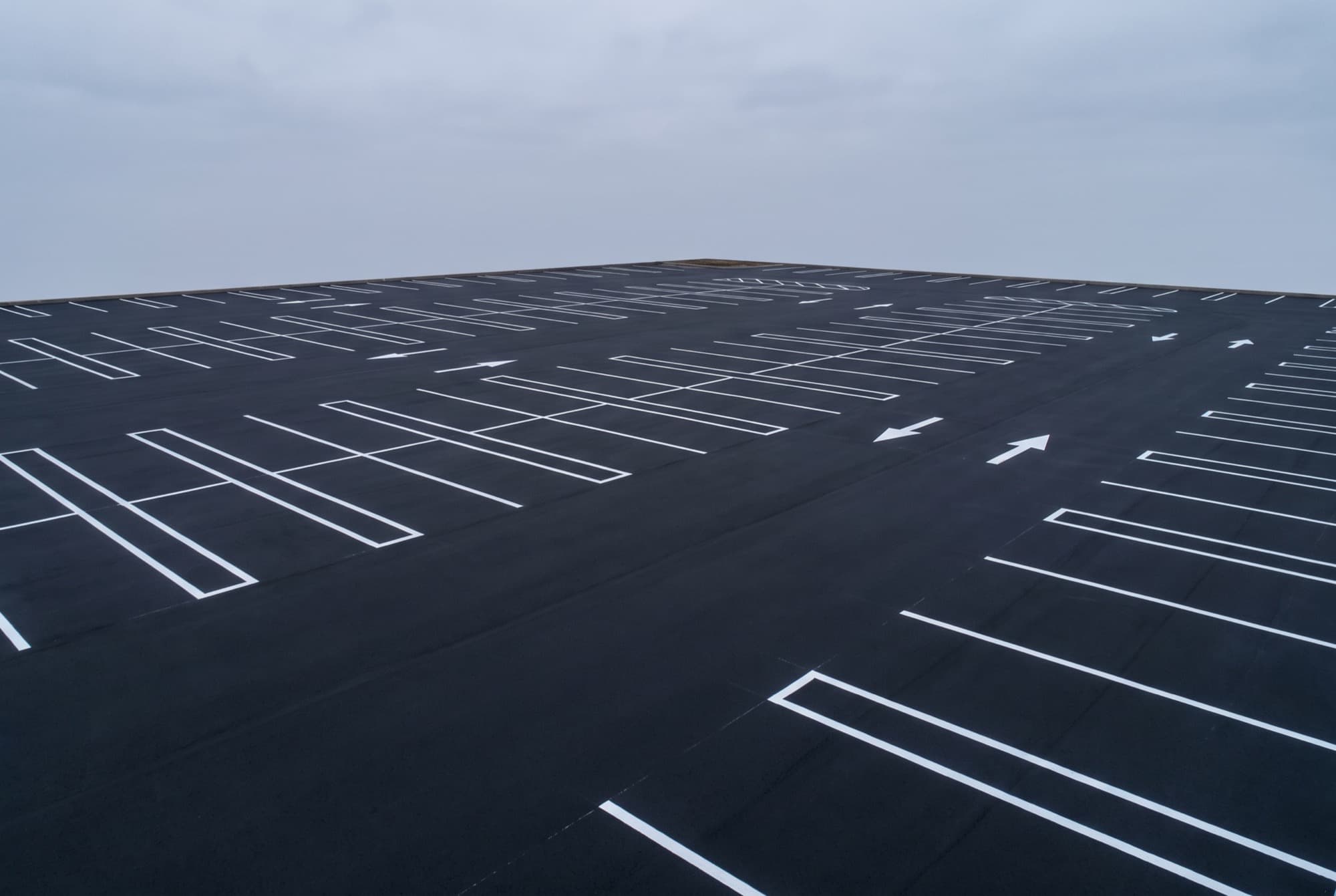An empty commercial parking lot the morning after it was sealcoated. The surface is a uniform matte black with no sheen or patchiness, and freshly repainted white stall lines and directional arrows sit crisply on top of it.